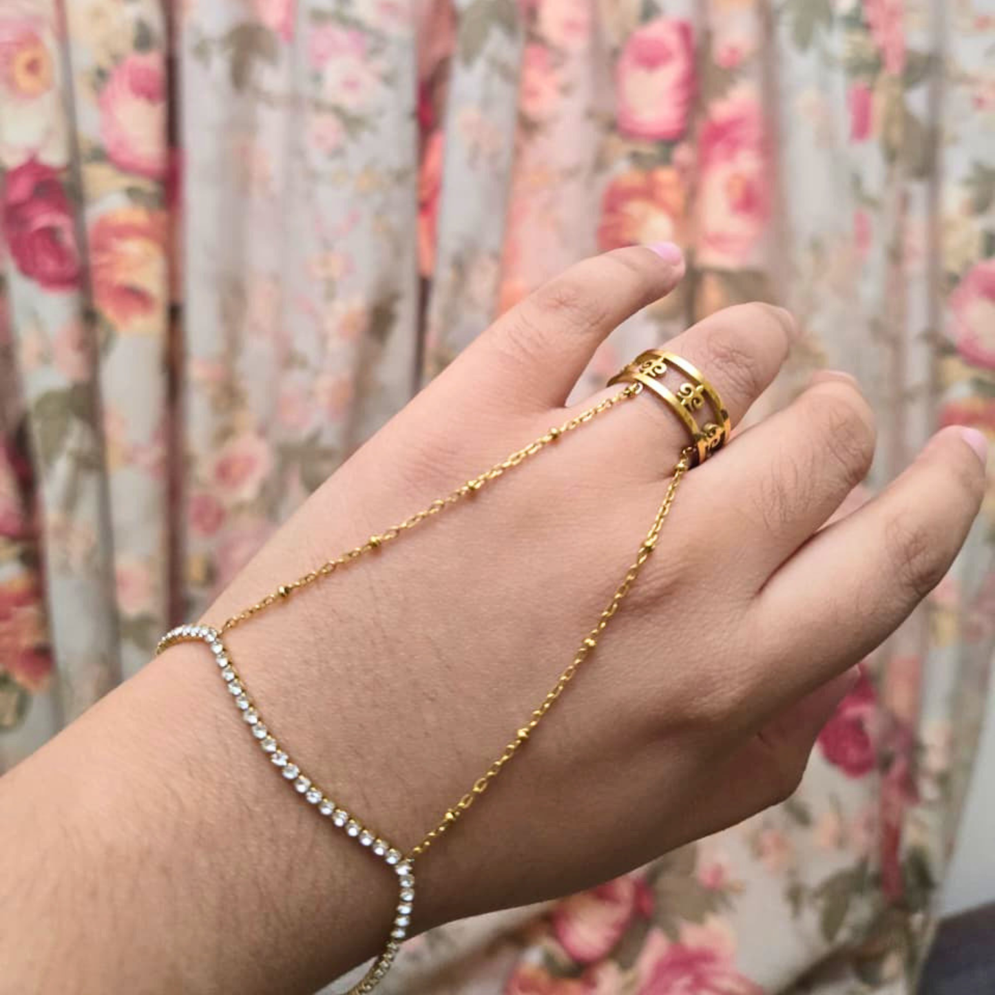 Hand wearing a gold bracelet and ring with a floral curtain background