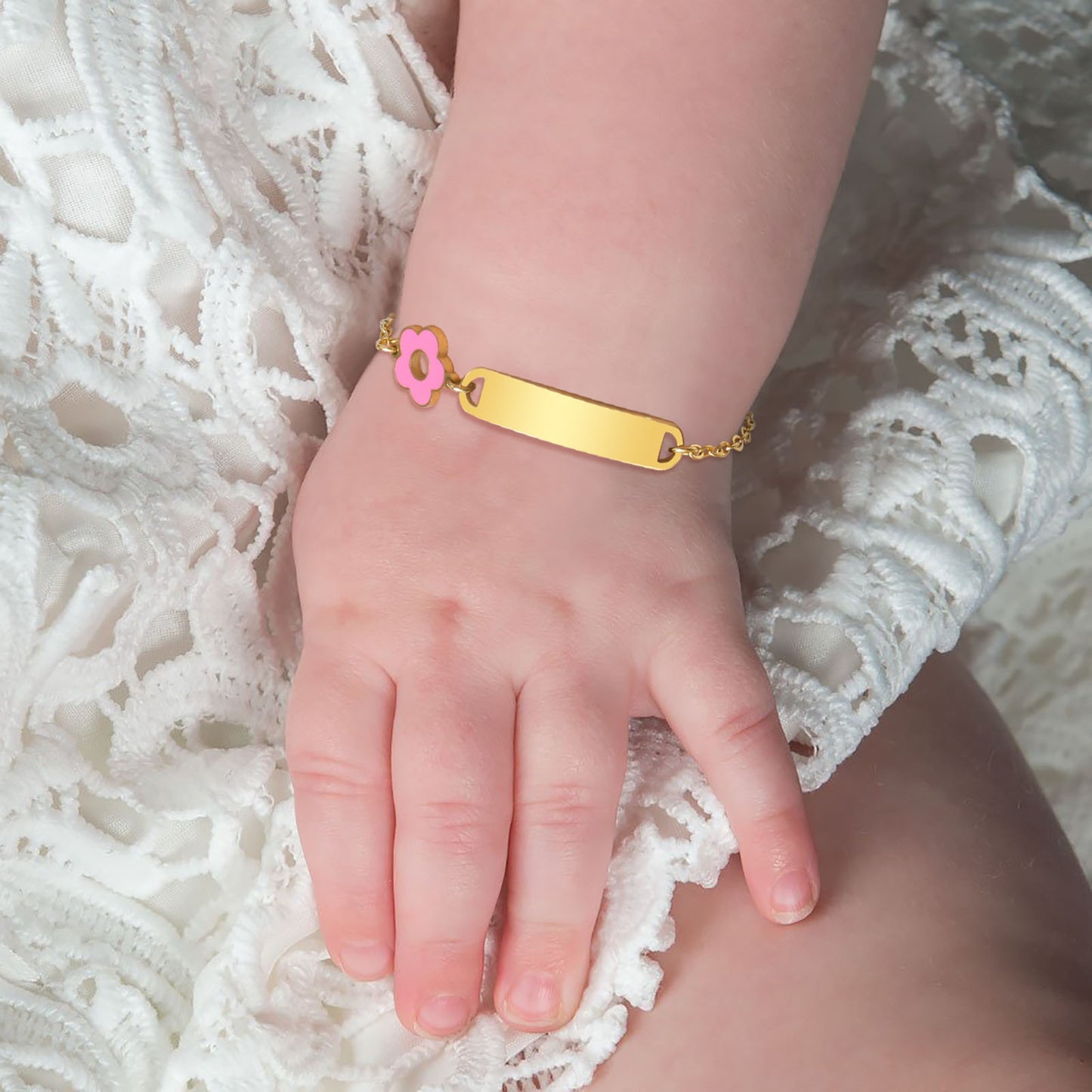 Gold baby bracelet with a pink flower charm on a child's wrist against a white lace background