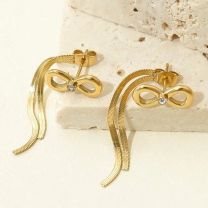 Gold bow-shaped earrings with chain details on a textured beige surface