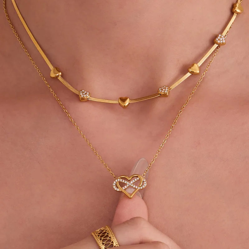 Gold necklaces with heart and infinity design on a person's neck against a pink background