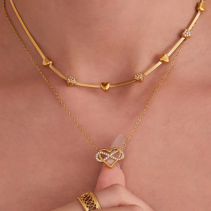 Gold necklaces with heart and infinity design on a person's neck against a pink background