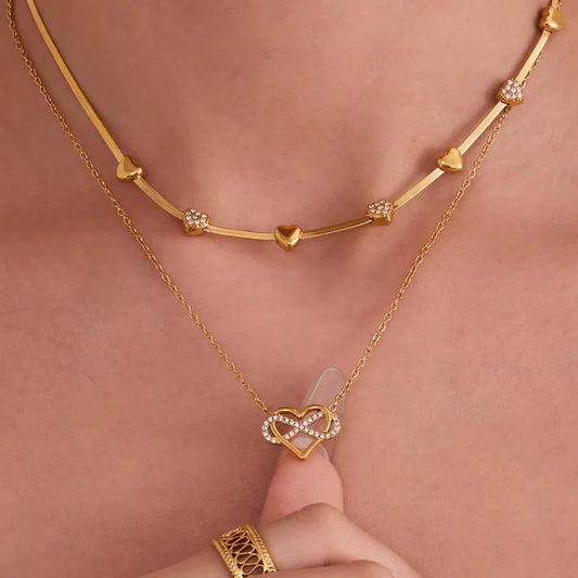 Gold necklaces with heart and infinity design on a person's neck against a pink background