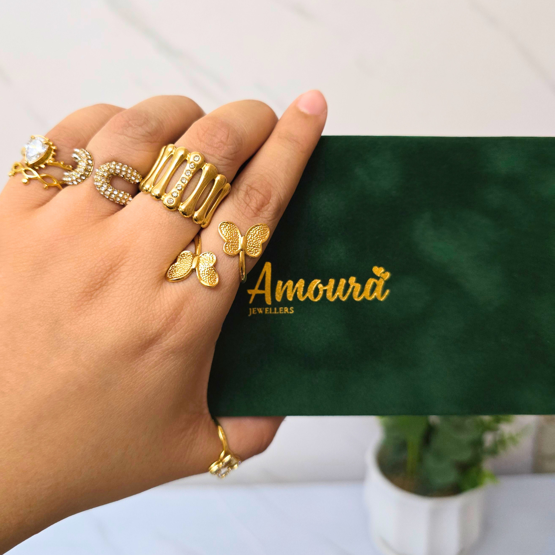 Hand wearing gold rings with a green box labeled 'Amoura Jewellers' in the background.