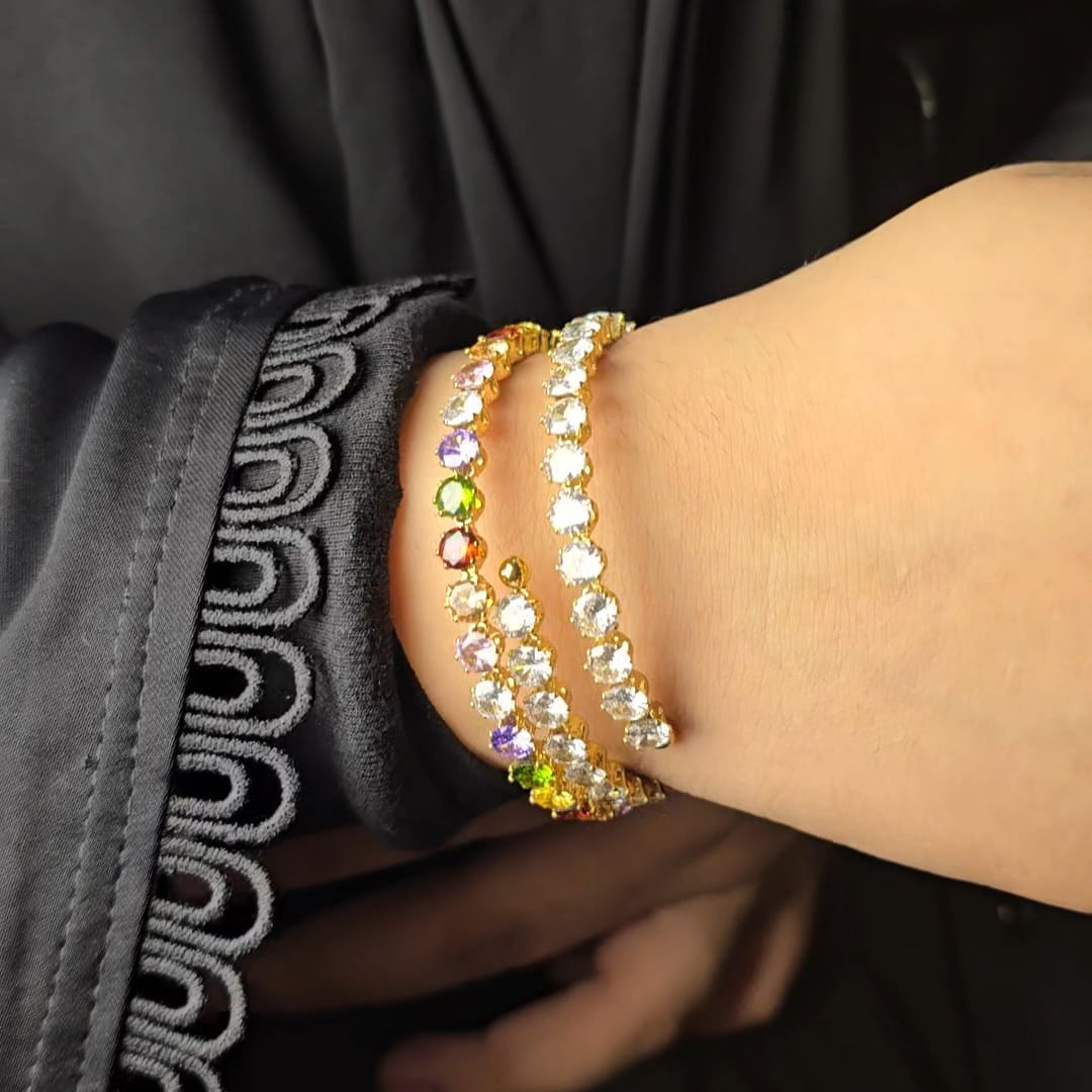 A close-up of a person's wrist wearing three stacked bracelets. The bracelets are gold-toned and set with clear round stones, with two of the bracelets featuring small multicolored accent stones.