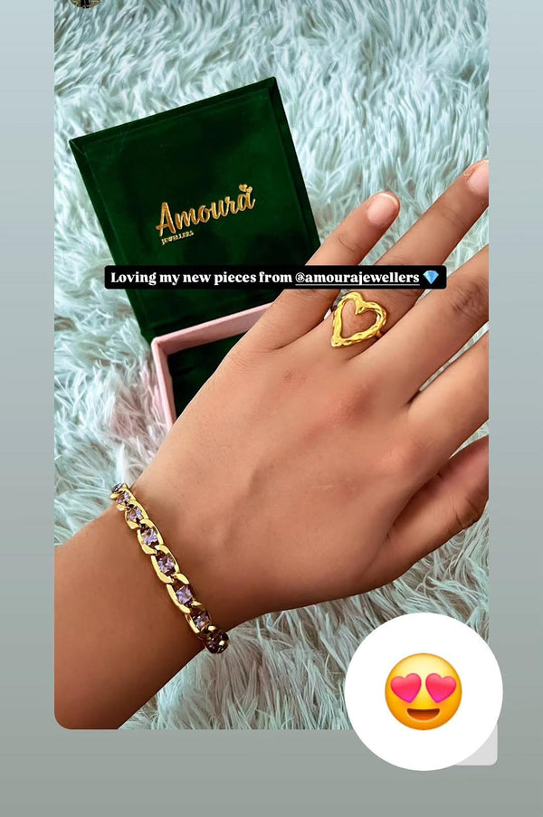 Hand wearing a gold bracelet and ring with 'Amoura Jewellers' jewelry box on a textured surface