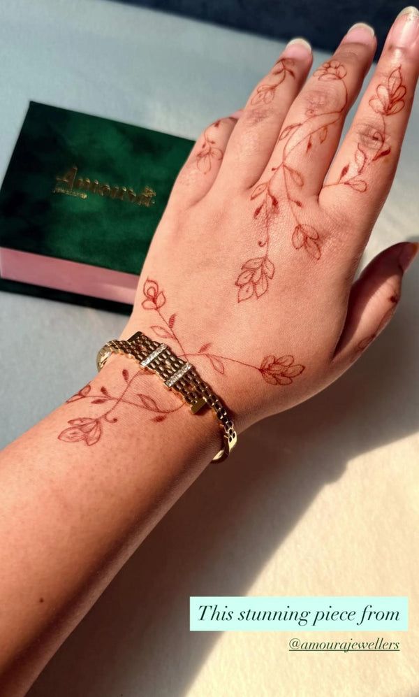 Hand with intricate henna design on wrist and palm, wearing a gold bracelet, with a blurred background.