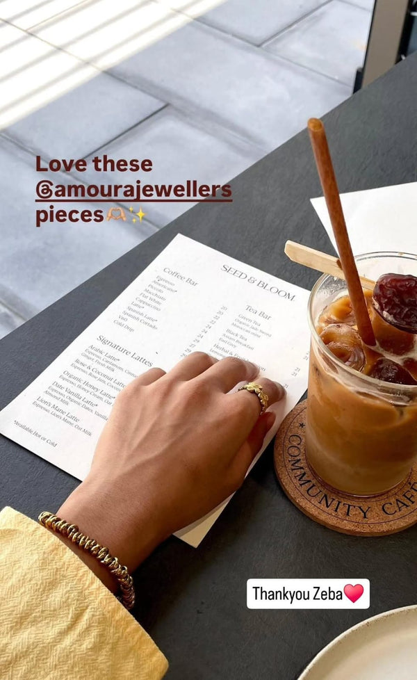 Hand holding a menu with a drink and jewelry, featuring 'Amourajewellers' on a table.