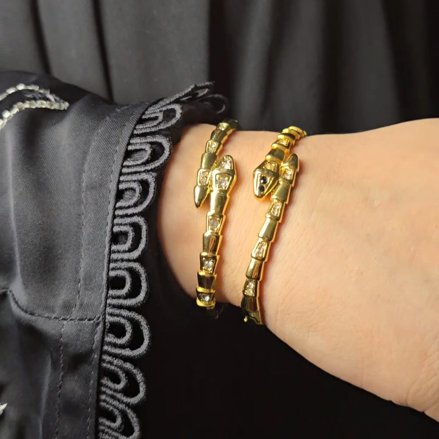 Two gold snake-themed bangle bracelets worn on a person's wrist, shown against a black garment with decorative gray embroidery.