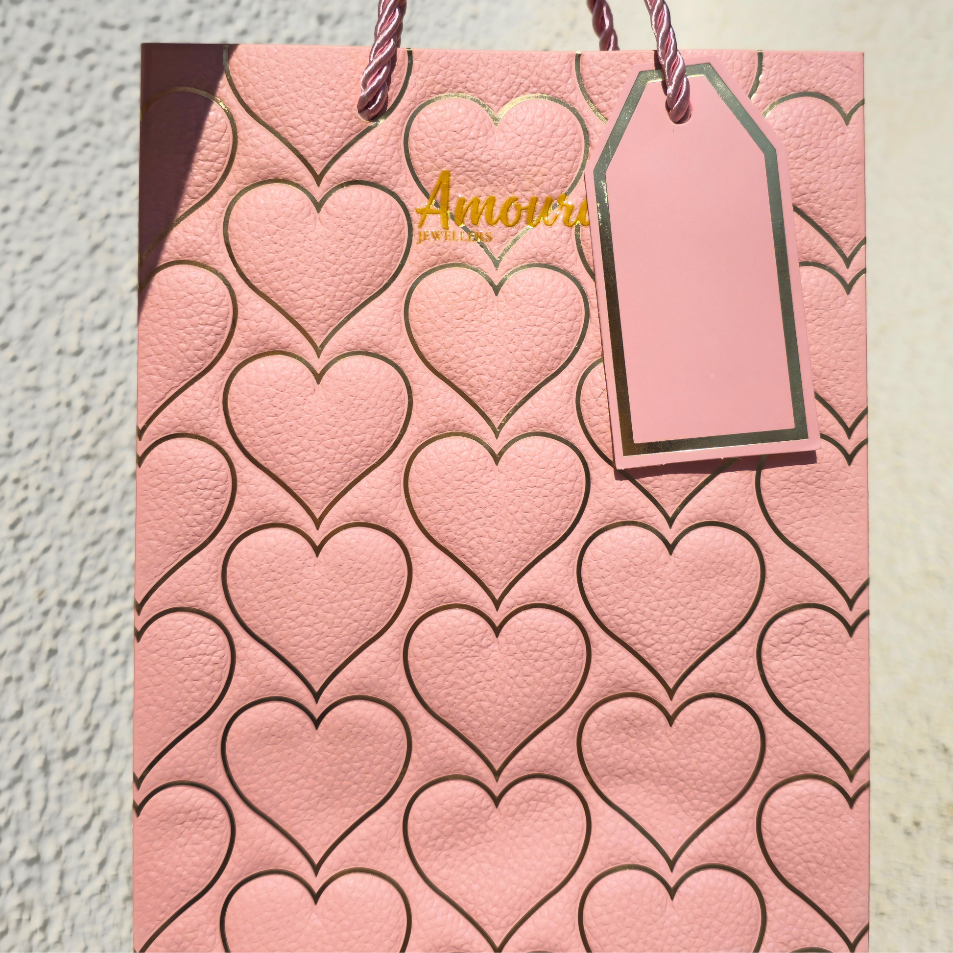 Pink gift bag with heart pattern and 'Amoura Jewellers' tag on a textured surface