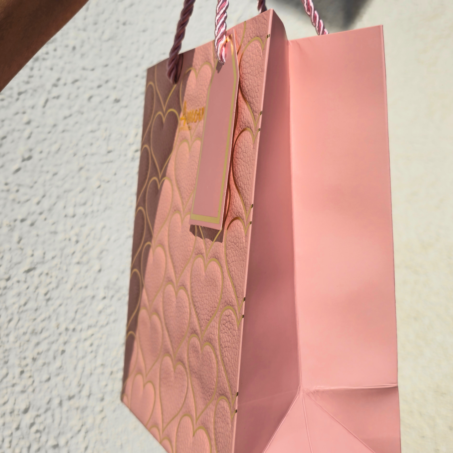 Pink gift bag with decorative pattern and tag against a textured wall.