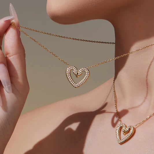 Gold heart-shaped necklace being worn and held by a hand against a neutral background