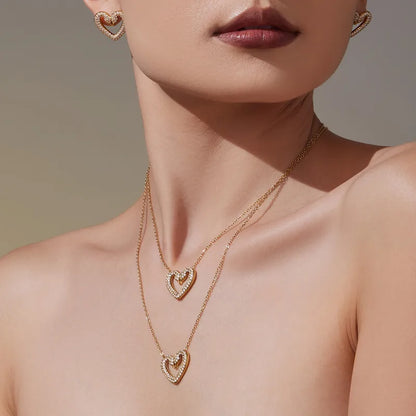 Close-up of a woman wearing a gold necklace and earrings with heart designs on a neutral background.